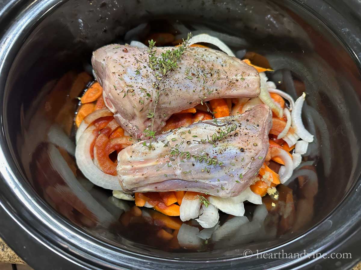 Slow cooker with raw lamb shanks seasoned on top of vegetables and a few sprigs of fresh thyme on top.