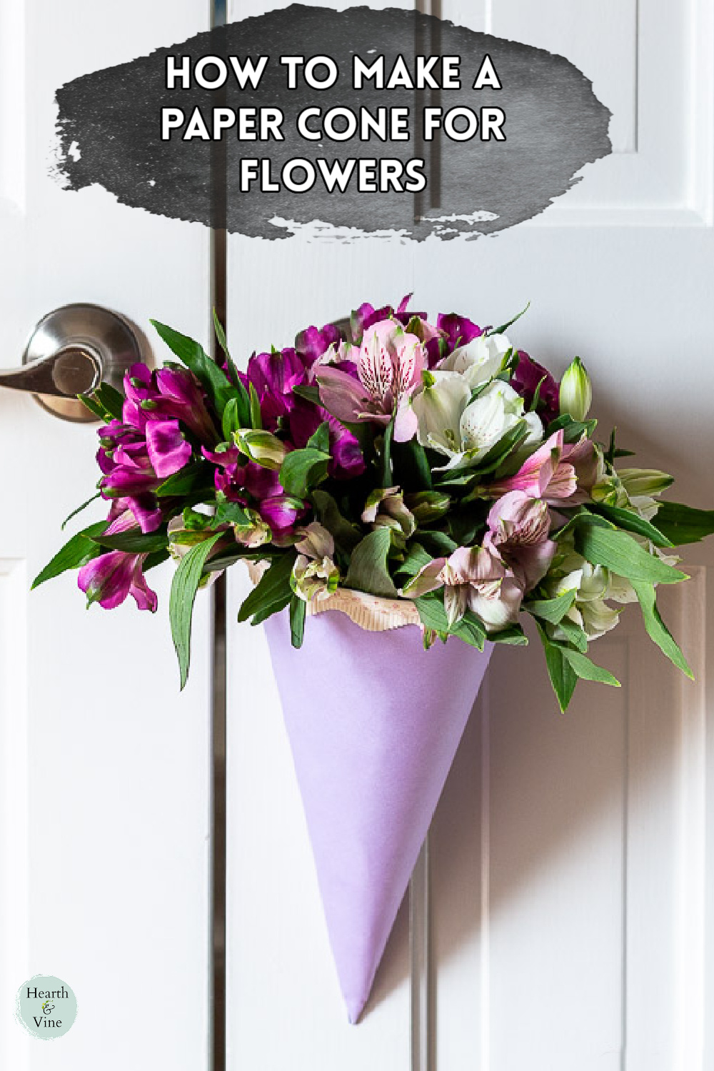 A lavender paper cone with pink ribbon edging holding a large bouquet of flowers hanging on a white door.