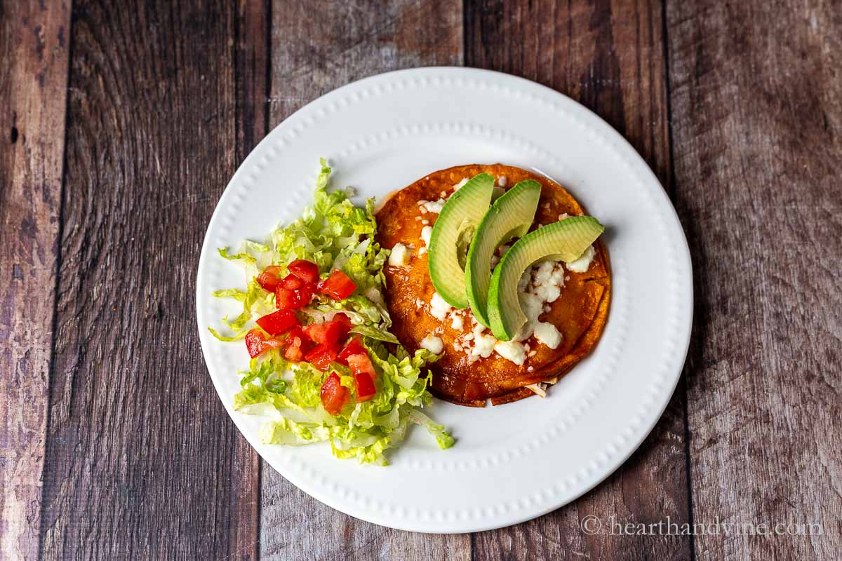 A flat enchilada with a side of shredded lettuce and chopped tomatoes and a few sliced of avocado on top.