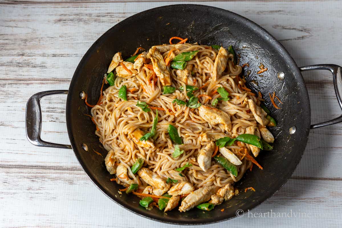 Large skillet with rice noodles and peanut sauce including chicken, carrots and snap peas.