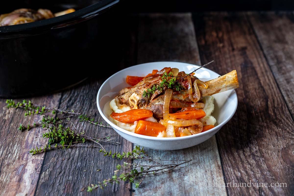A serving bowl with mashed potatoes topped with a slow cooked lamb shank, carrots, onions and thyme.