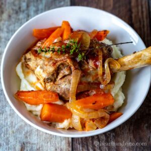 Small bowl with mashed potatoes topped with a slow cooked lamb shank, carrots and onions.