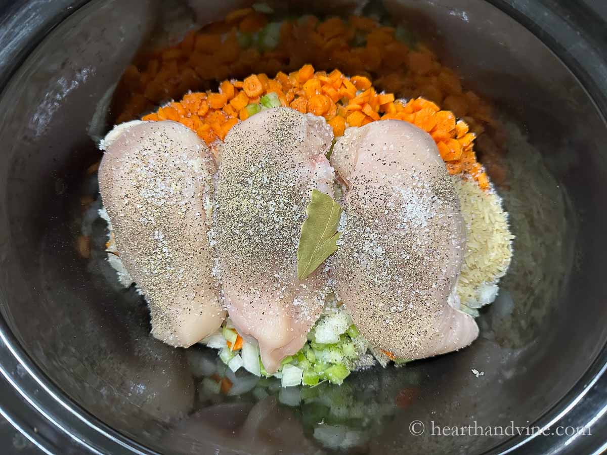 Slow cooker with carrots, celery, onoins and rice. Three boneless, skinless chicken breasts on top with salt and pepper and a bay leaf.