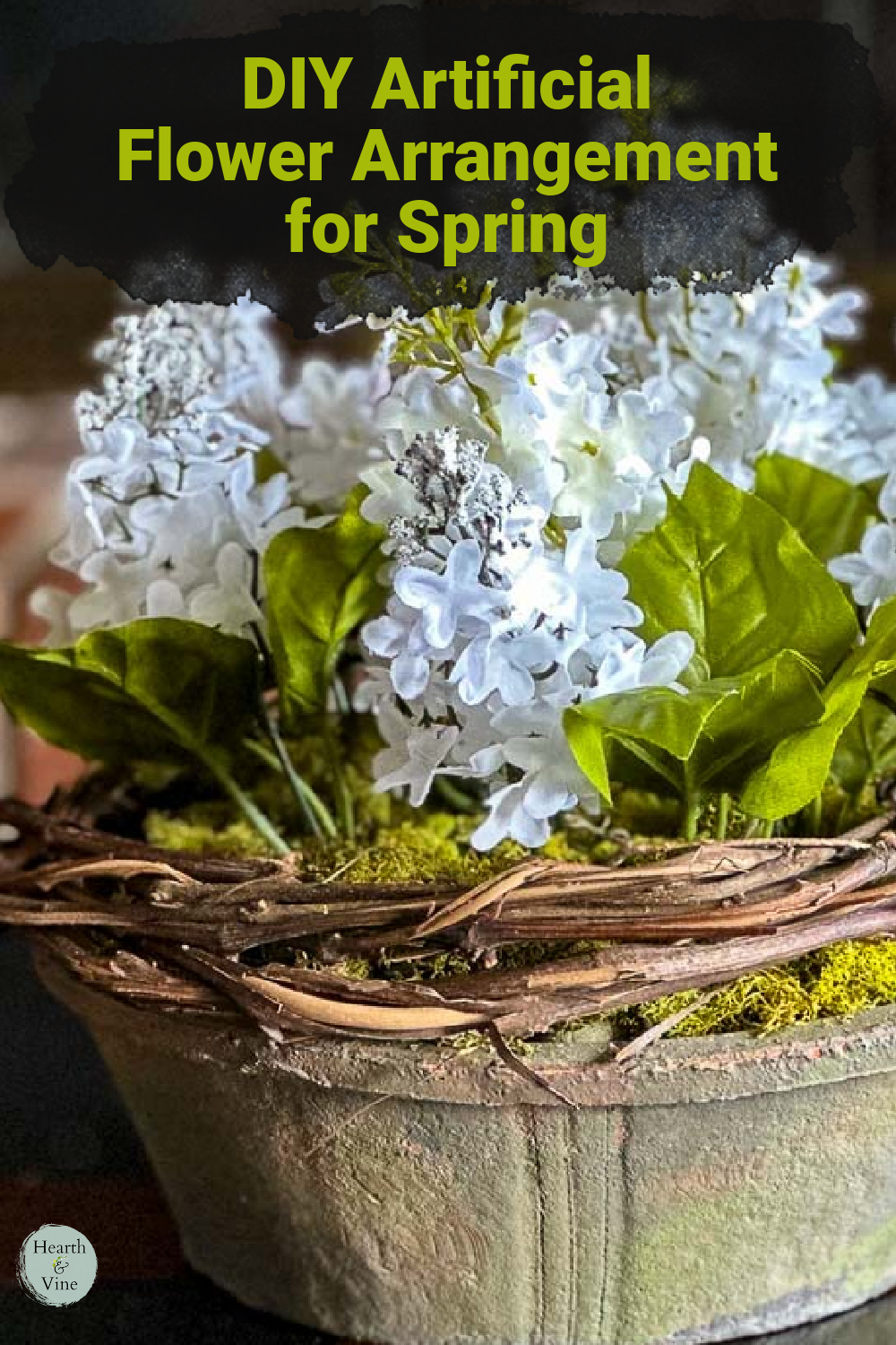 A close up image of an artificial flower arrangement with white lilacs, moss, grapevine in a clay pot.