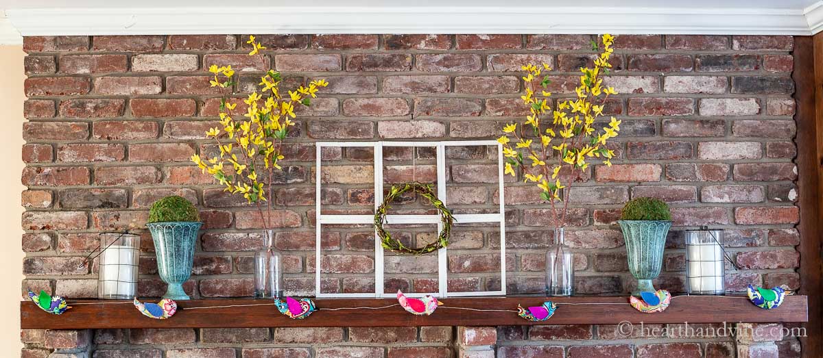 Spring mantel with farmhouse window frame and wreath in the center followed by forsythia blooms, moss balls and candles. A fabric bird garland hangs below in front.