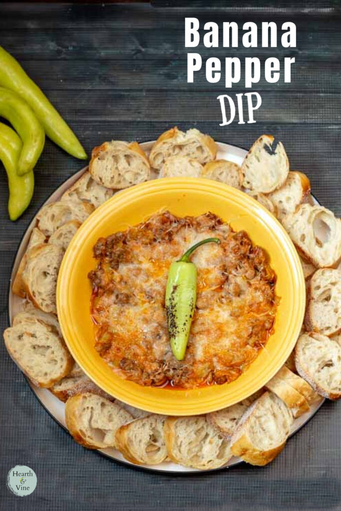 Bowl of stuffed banana pepper dip surrounded by sliced baguette bread. A charred banana pepper is set on top for garnish.