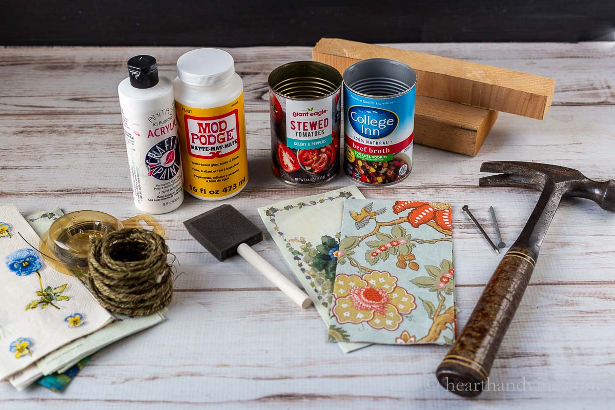 Supplies for the tin can baskets including Mod Podge, white paint, a hammer, paper napkins, two tin cans, wire and a sponge brush.