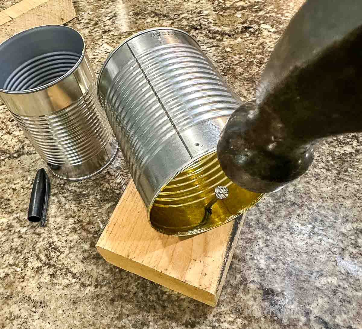 A large nail being hammered into the side of a can to make a hole.