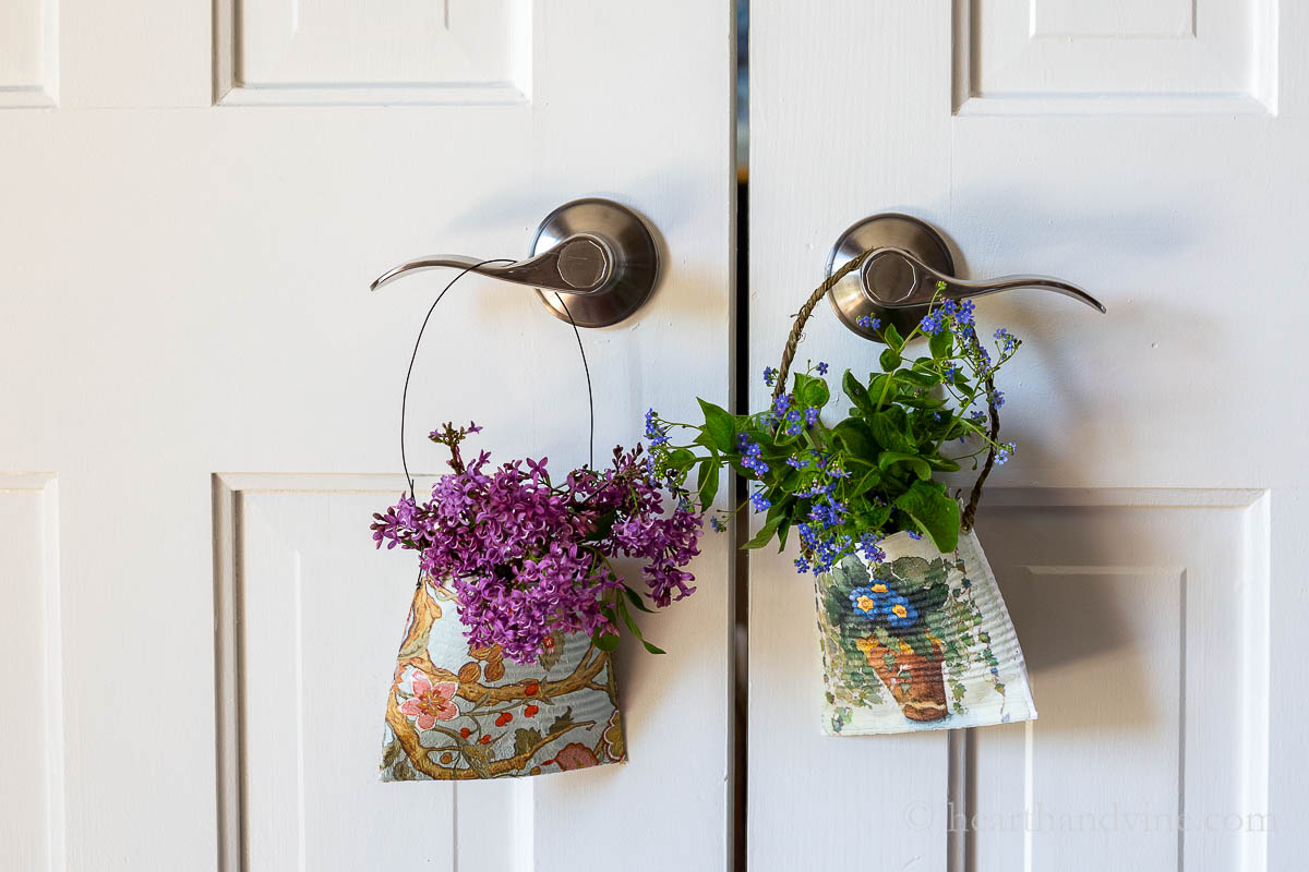 Two smashed cans hanging on door knobs. One with lilacs and the other with blue flowers.