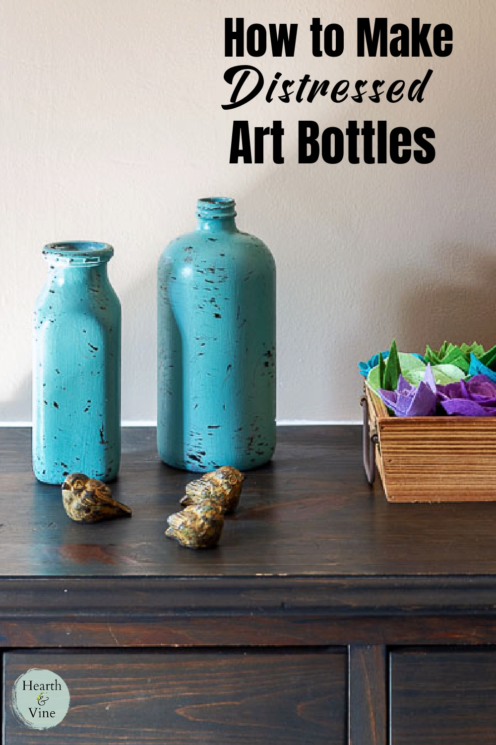 Two blue aged bottles on a table with little birds in front and a box of felt succulents to the side.