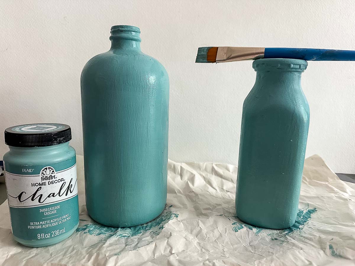 A finishing coat of blue paint on two bottles with a bristle brush.