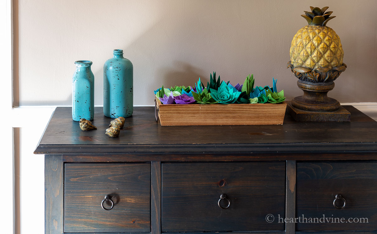 Two painted glass bottles on a chest in the foyer with tiny birds, a box of felt succulents and a large resin pineapple.