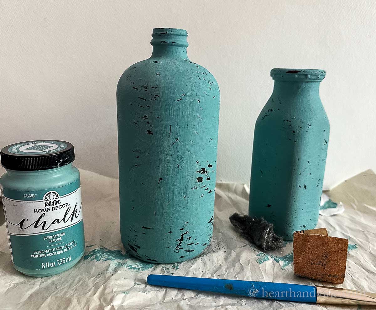 Two distressed painted bottles with sandpaper and a jar of chalk paint.