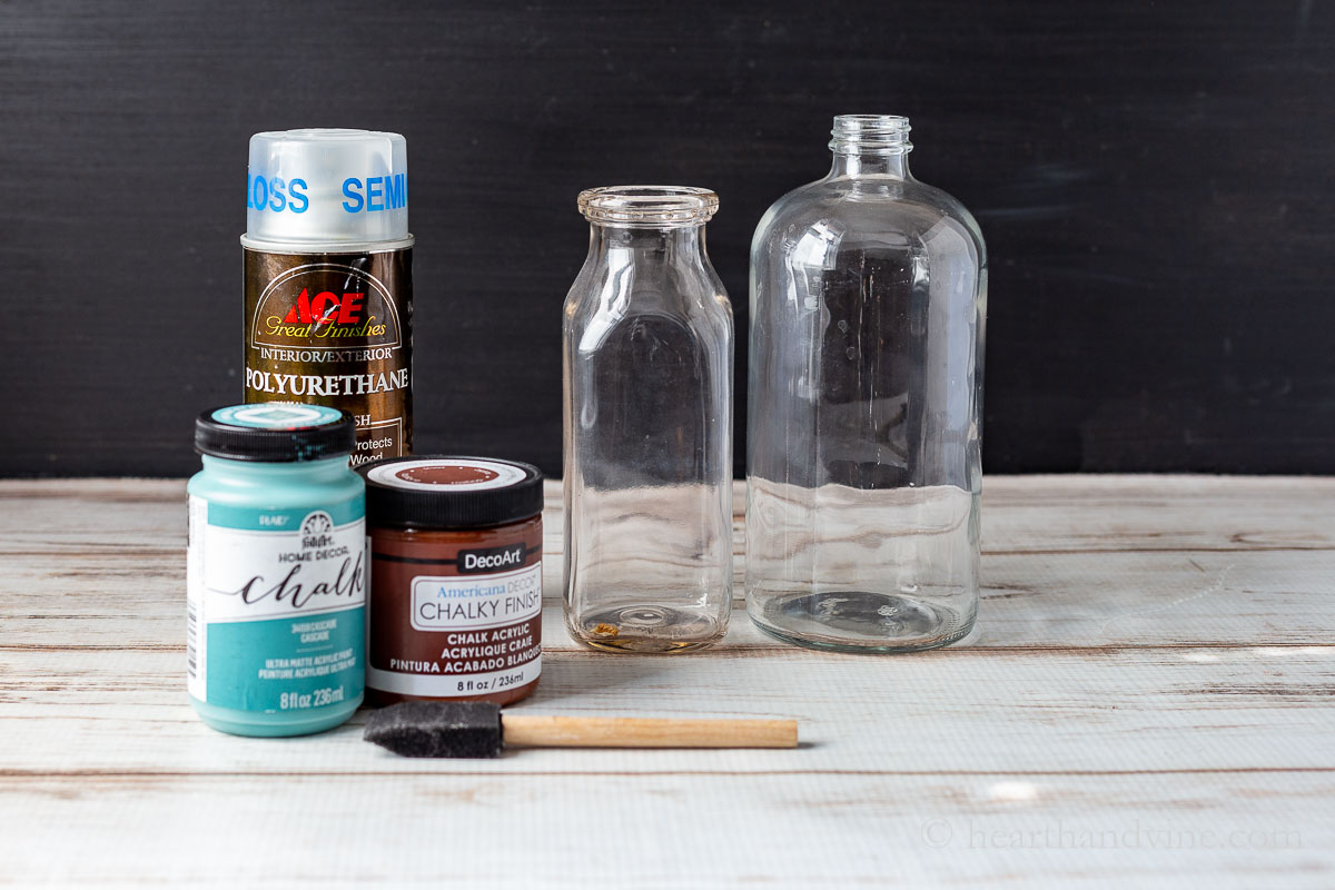 Supplies for the painted bottle craft including two glass bottles, brown and blue chalk paint, poly spray and a sponge brush.