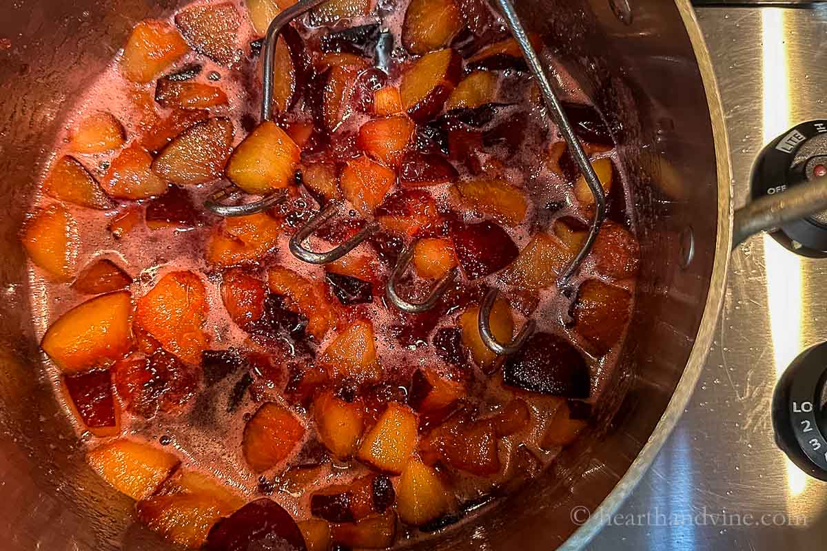 Plum mixture cooking down in a pot on the stove. A potato masher smashing the fruit.