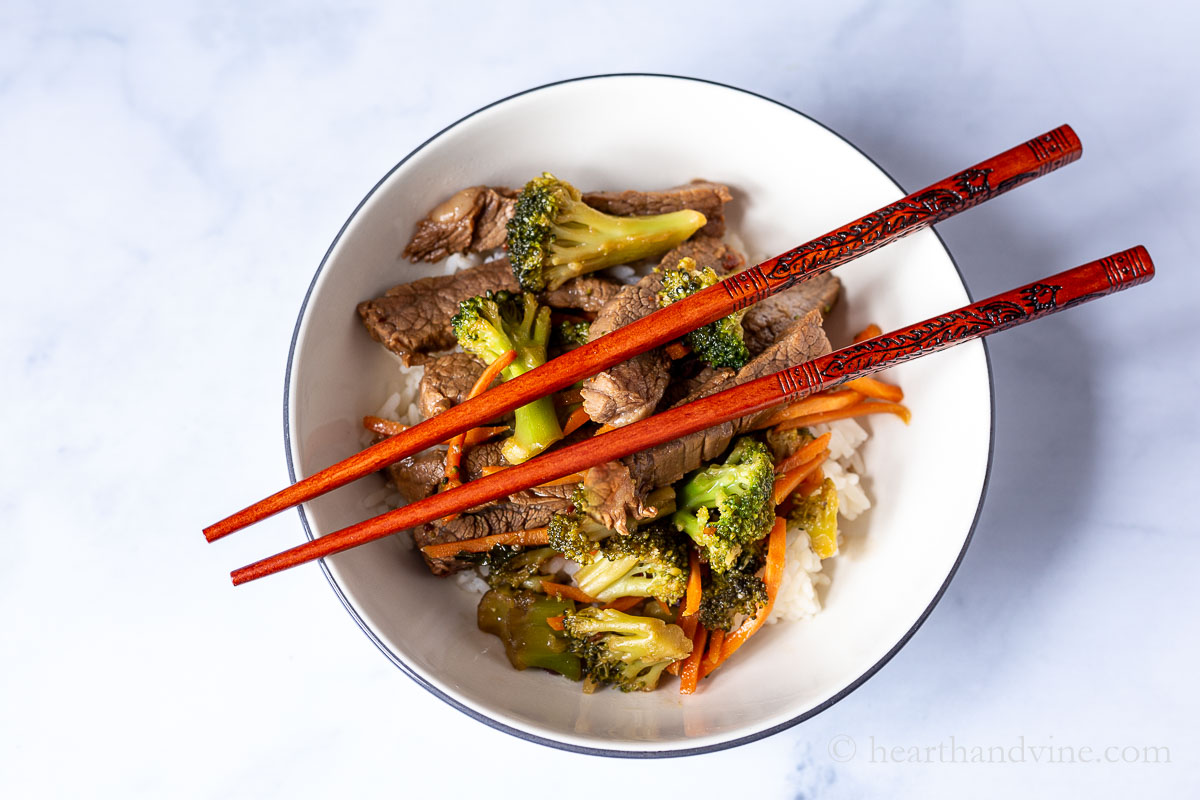 Stir fried beef and broccoli in a bowl over rice with chopsticks across the top.