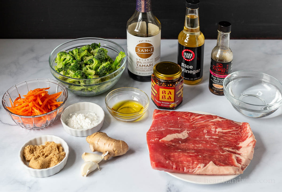 Ingredients for beef and broccoli including flank steak, broccoli florets, carrot sticks, tamari, rice vinegar, sesame oil, garlic, ginger, chili crisp, brown sugar and cornstarch.