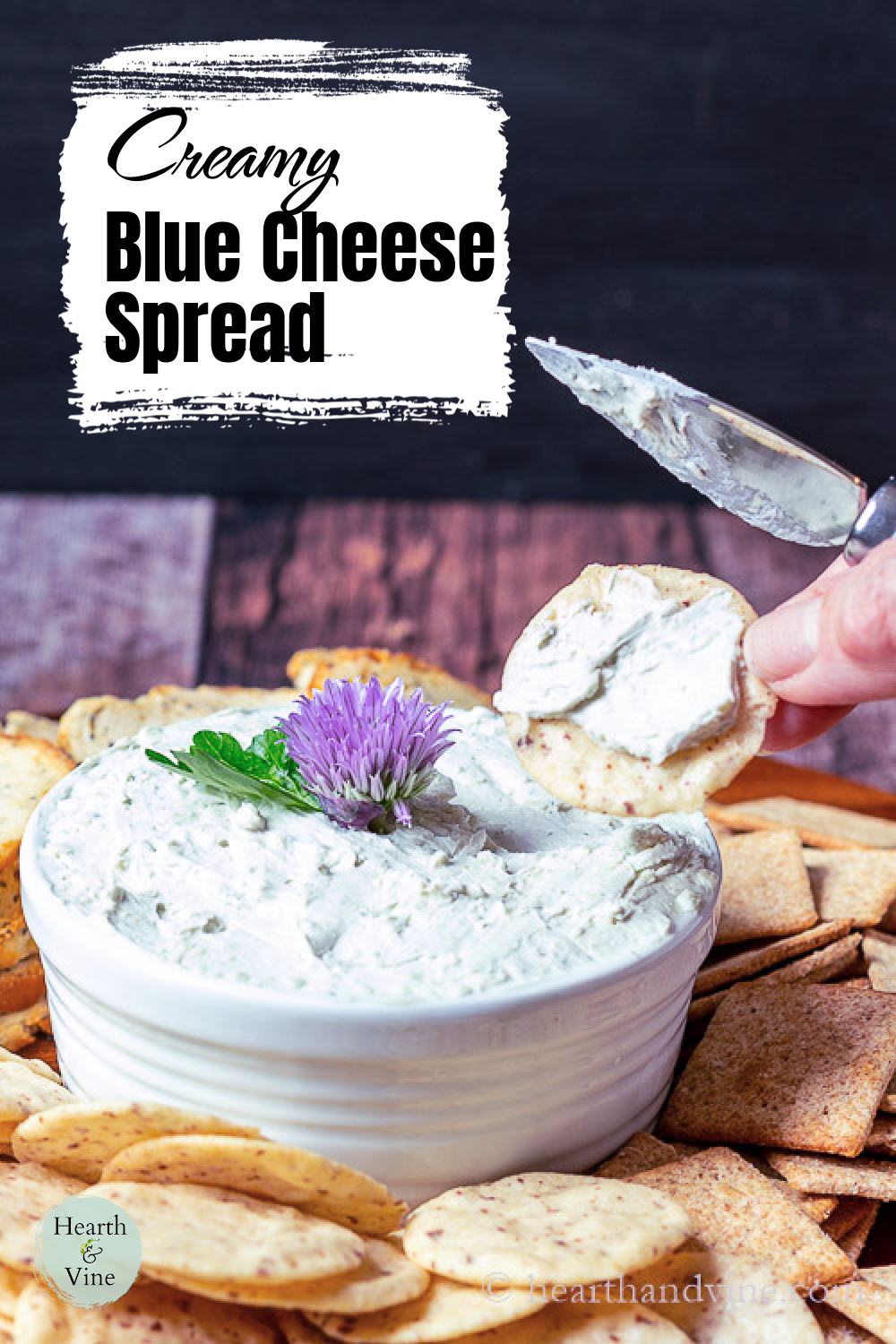 Cracker in hand with blue cheese spread next to a small bowl of the spread with a chive flower on top, surrounded by a few different types of crackers.