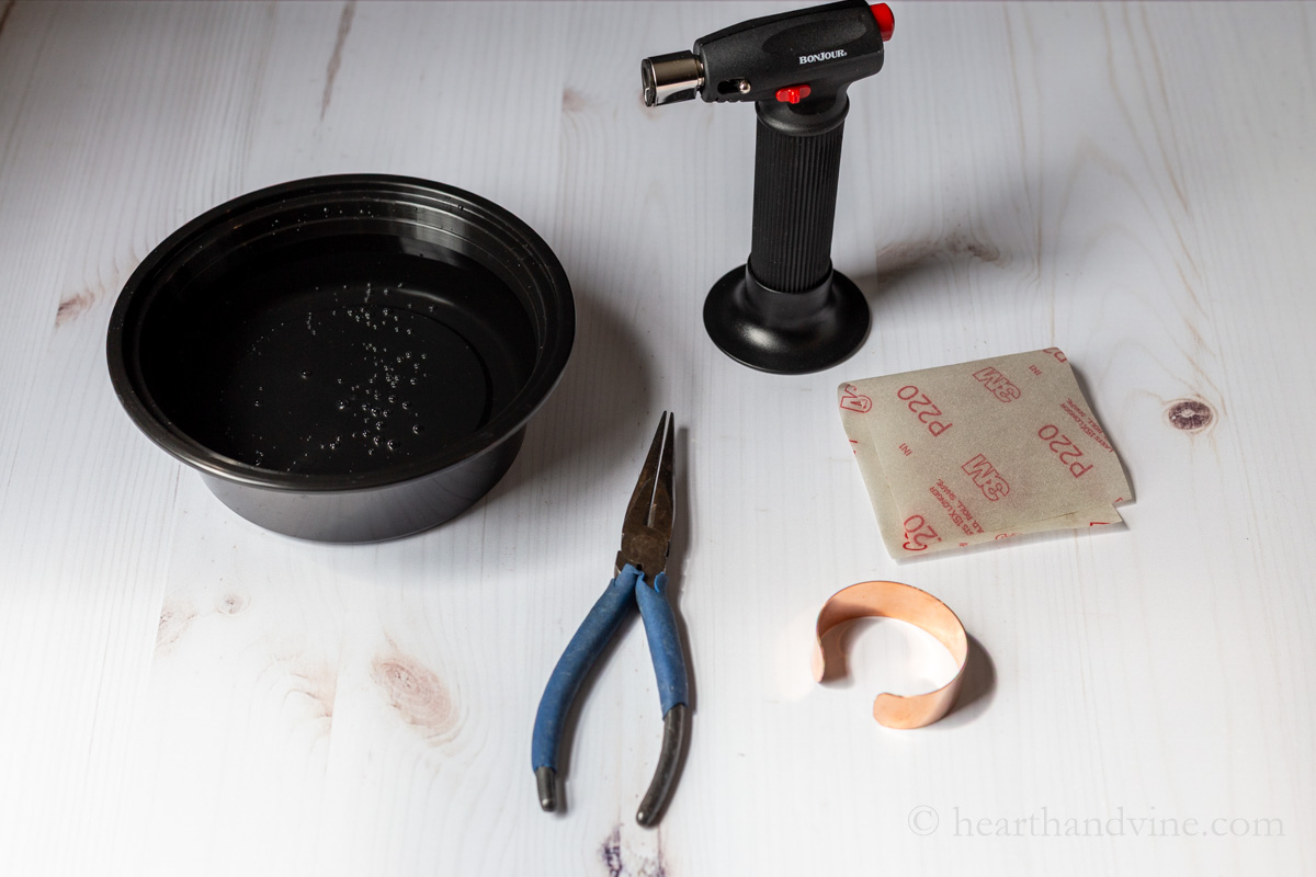 Supplies to flame paint a copper bracelet including needle nose pliers, sandpaper, a bowl of water and a kitchen torch.