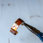 Needle nose pliers holding a flame painted copper bracelet.
