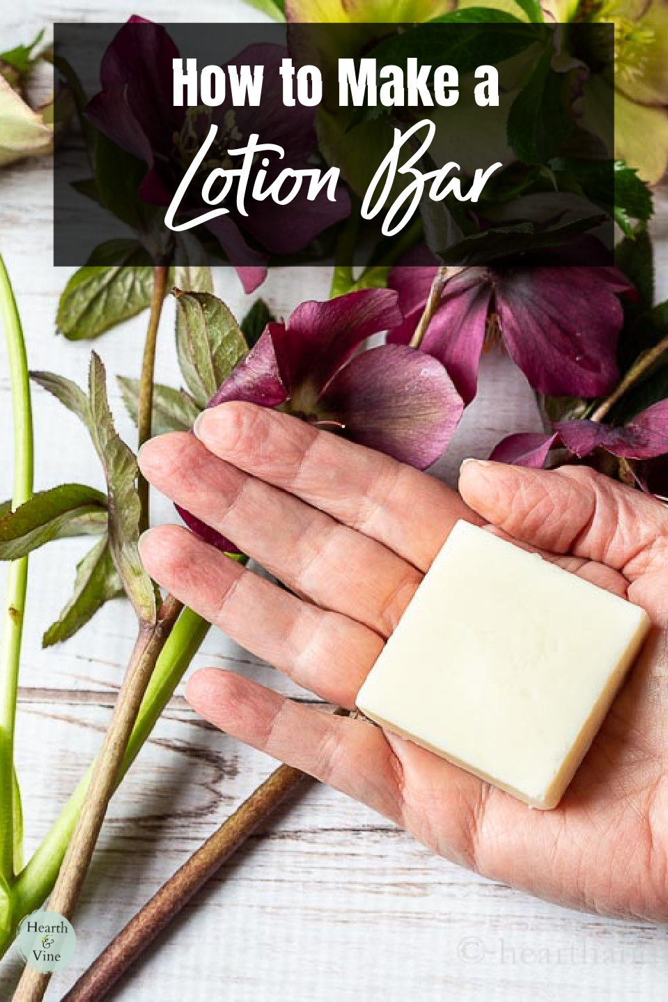 A hand holding a square lotion bar above some hellebore flowers.