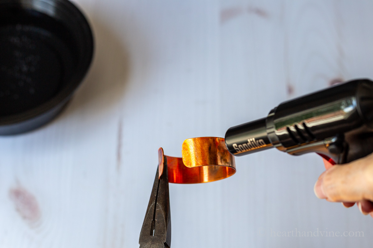 A kitchen torch heating a copper cuff that is held with pliers.
