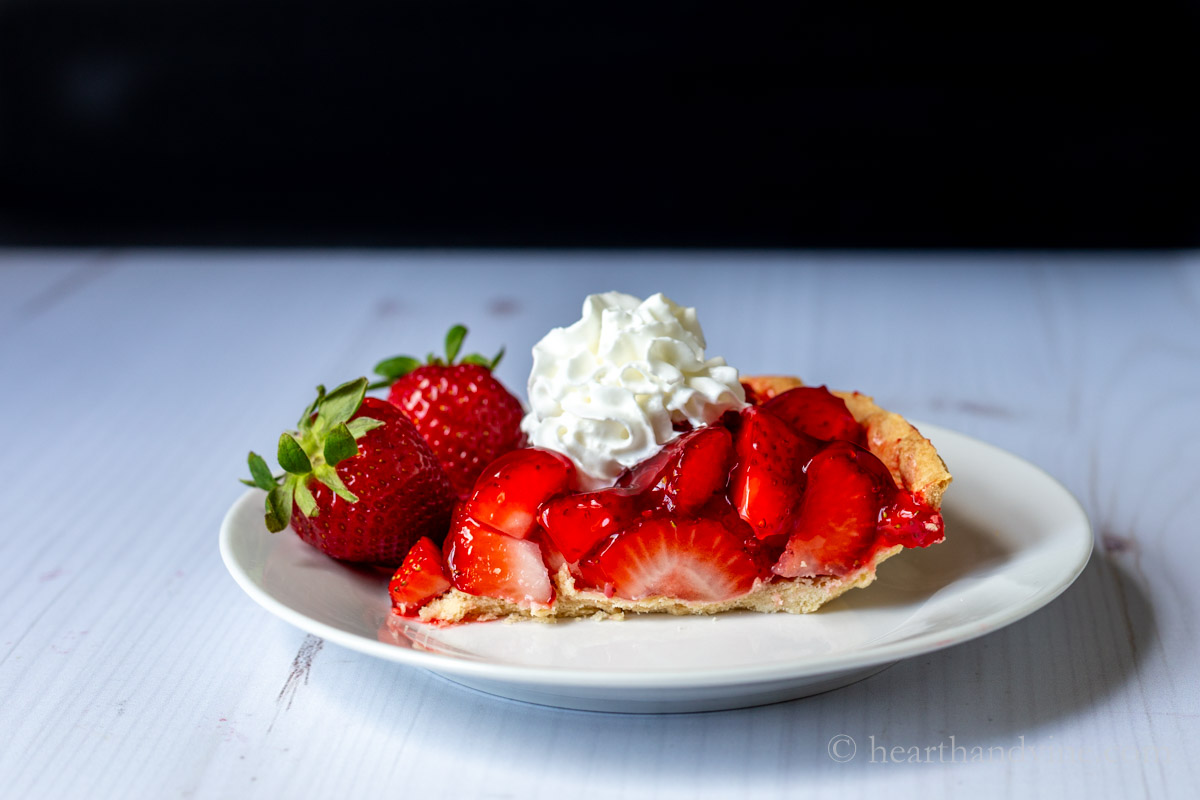 Side view of a slice of strawberry glazed pie with a dollop of whipped cream and a couple of whole berries on the side of the plate.