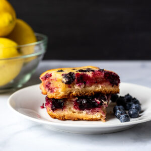 Two lemon blueberry bars stacked one a plate with lemons in the background and fresh blueberries on the plate.