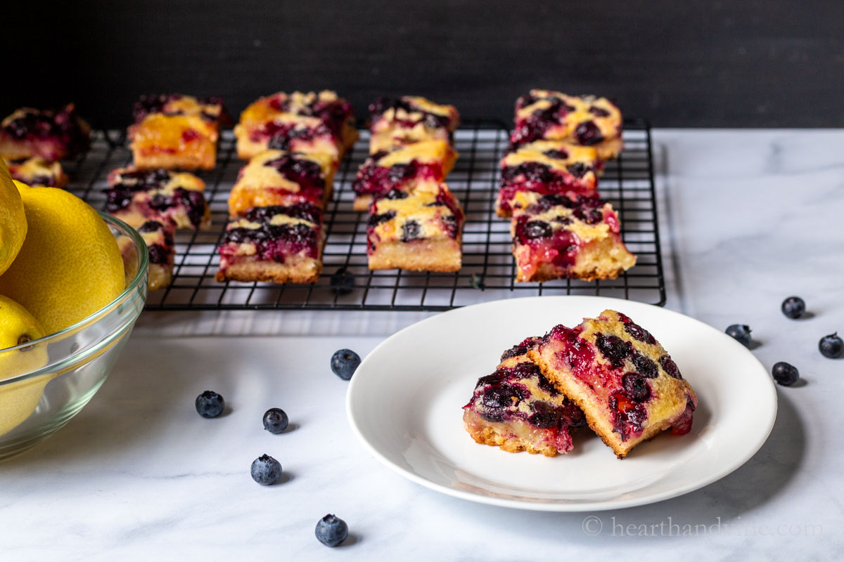 A couple of lemon blueberry bars on a plate with scattered fresh blueberries around and the rest of the bars on a cooling rack behind.