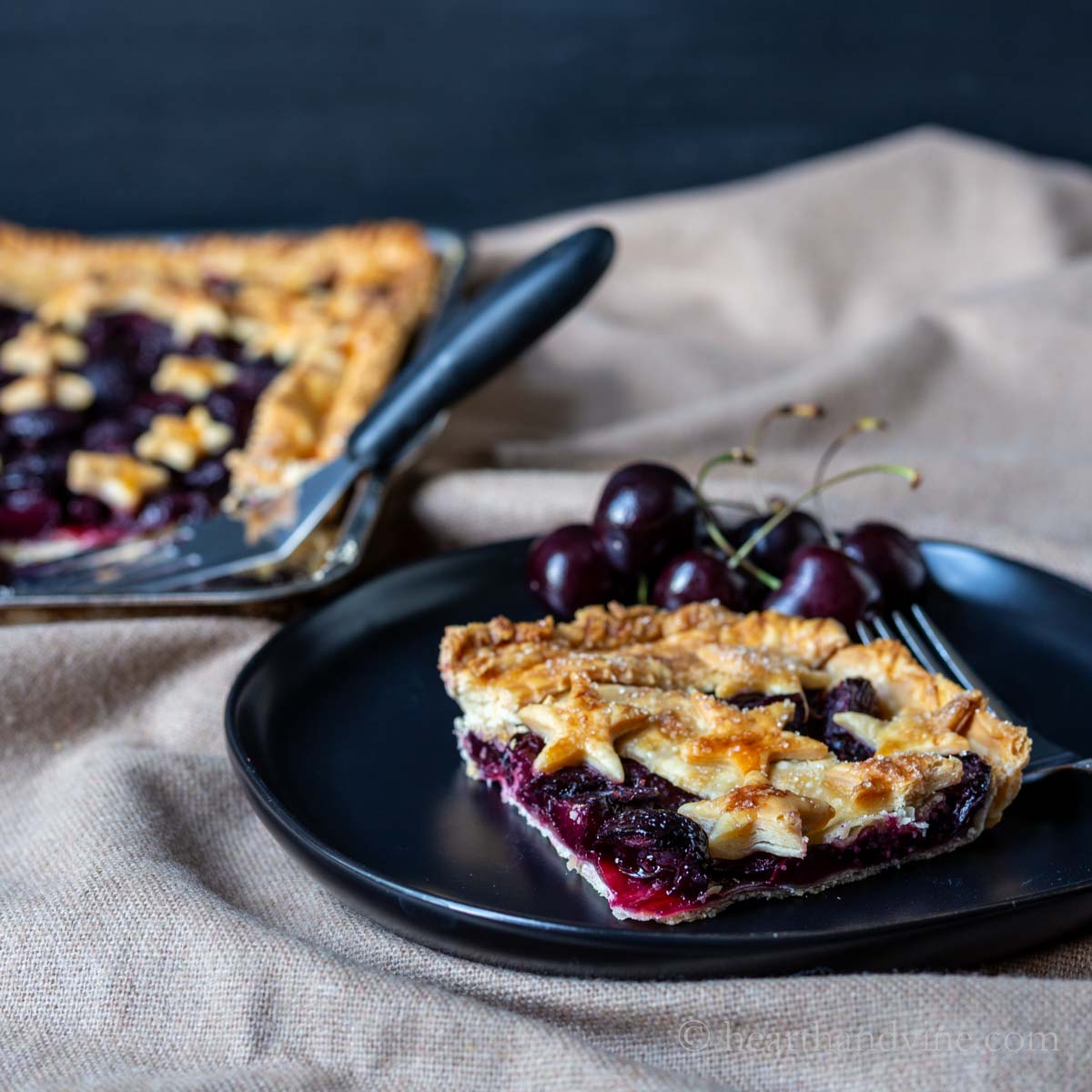 Corner slice of cherry slab pie on a black plate with a few fresh cherries and the entire slab in the background.