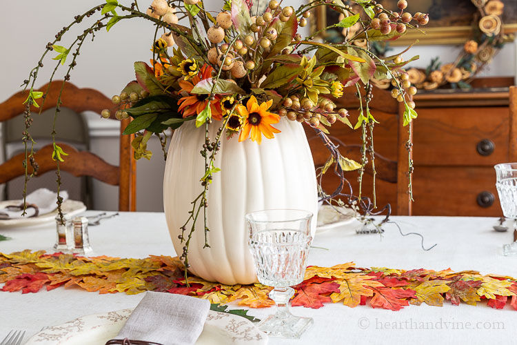 Large cream faux pumpkin with artificial flowers and berries arranged in the top for a pretty fall centerpiece on a faux autumn leaf runner.