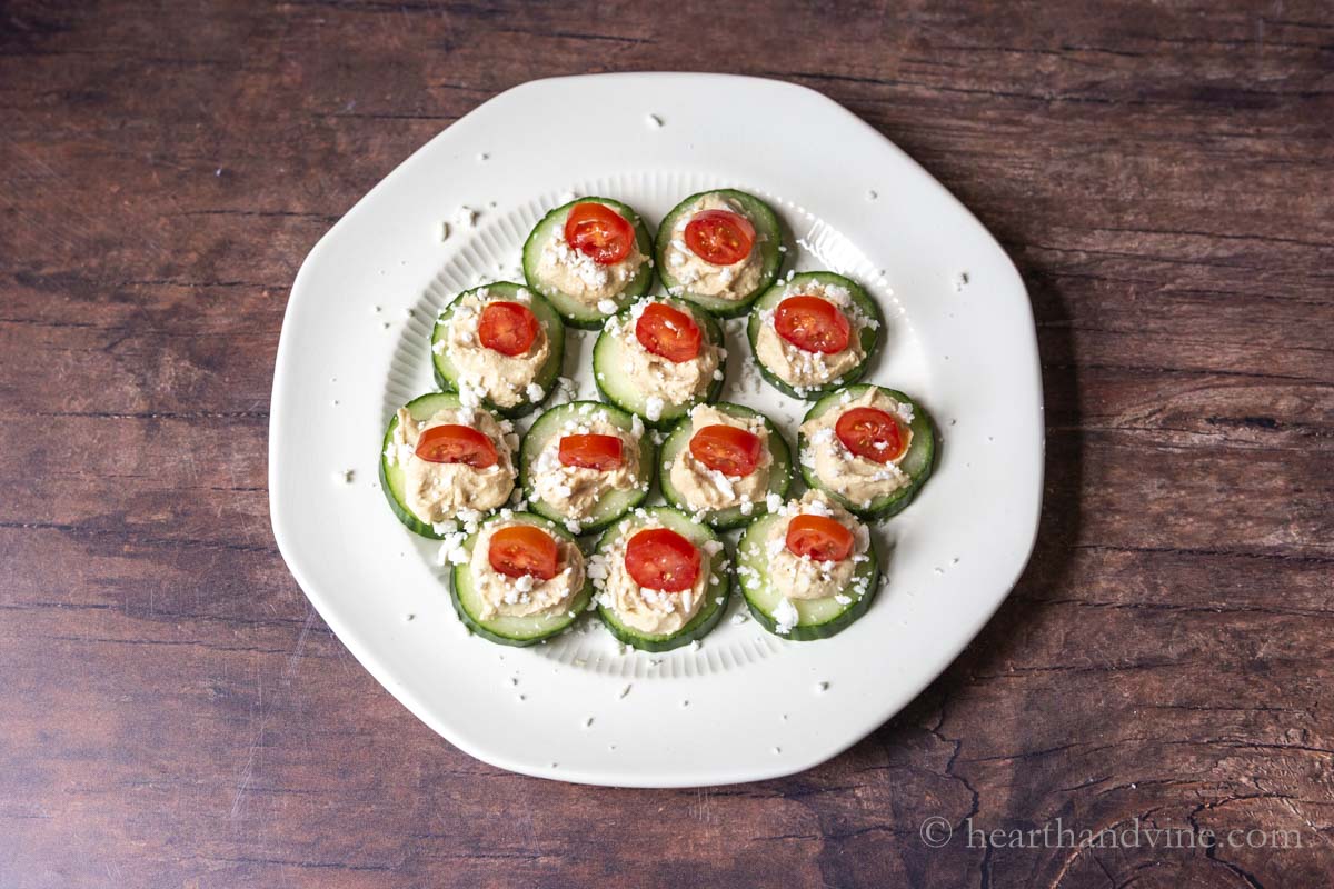 A white plate with cucumber rounds topped with hummus, feta cheese and a slice of tomato.