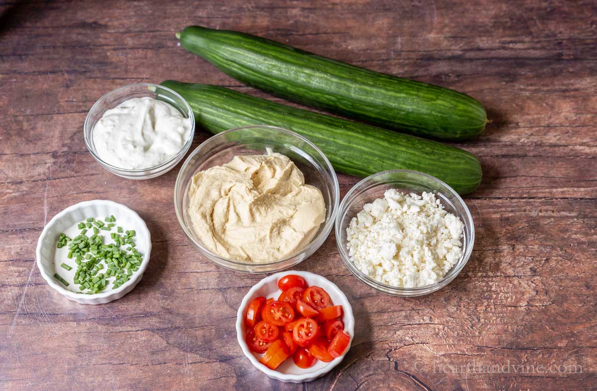 Two English cucumbers and bowls of ranch dip, chopped chives, hummus, crumbled feta and sliced tomatoes.
