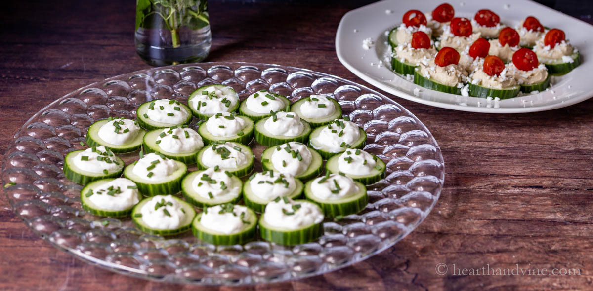 Cucumber bites with ranch and chives next to a Greek flavored cucumber bites with hummus, feta and tomatoes.