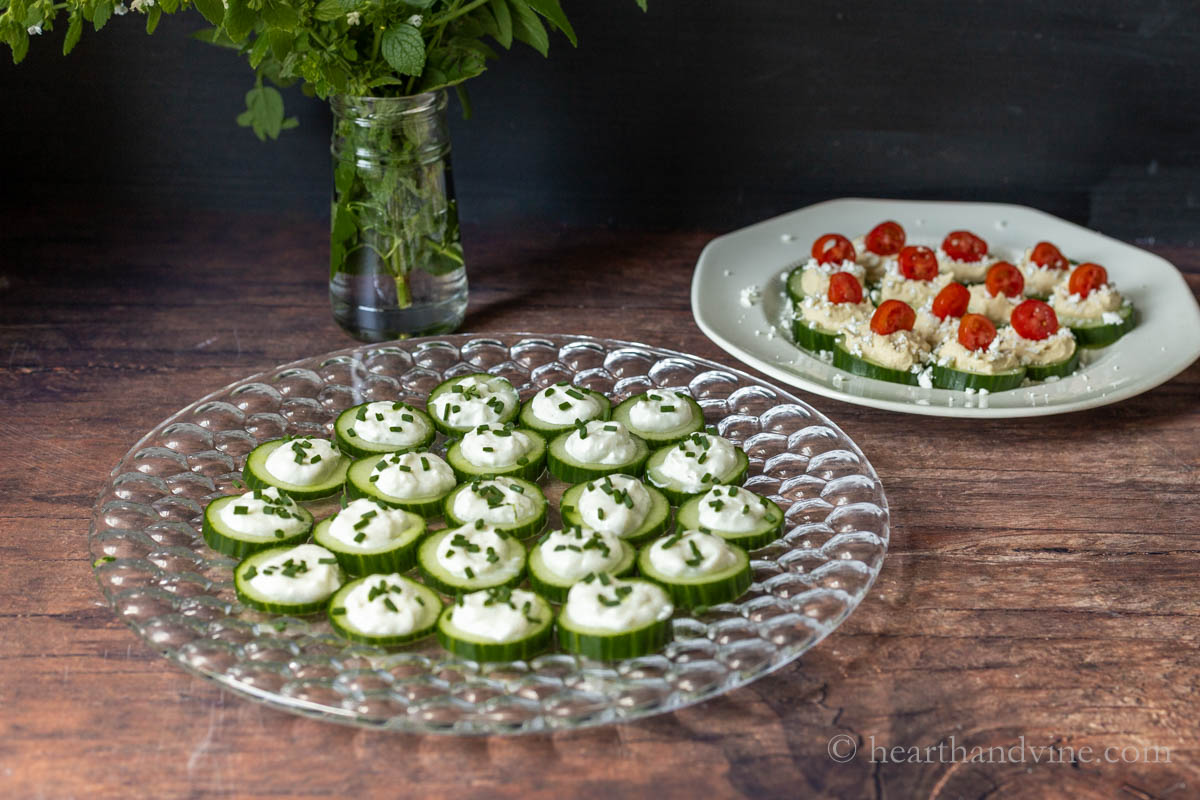 Tray of ranch and chive cucumber bites next to cucumber bite with hummus, feta and tomatoes.