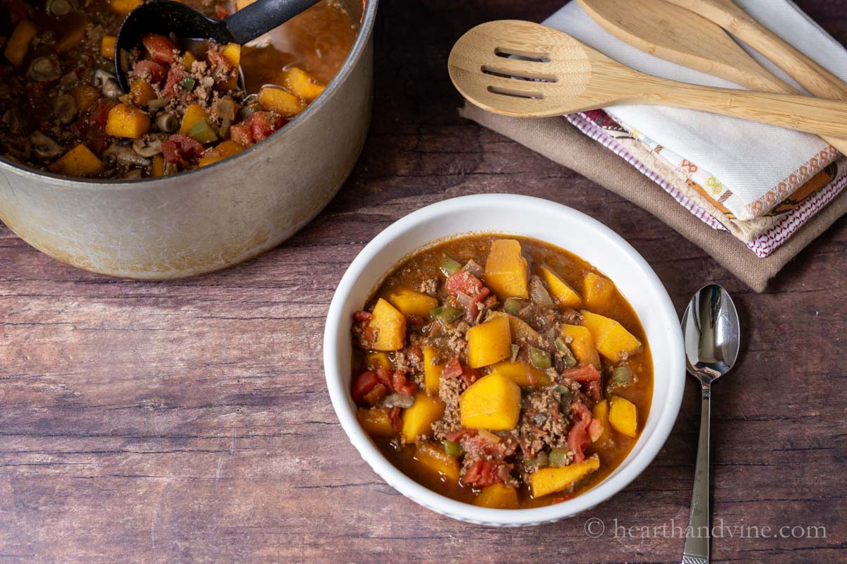 A bowl of pumpkin chili with a spoon on the side and an entire pot of chili in the background.