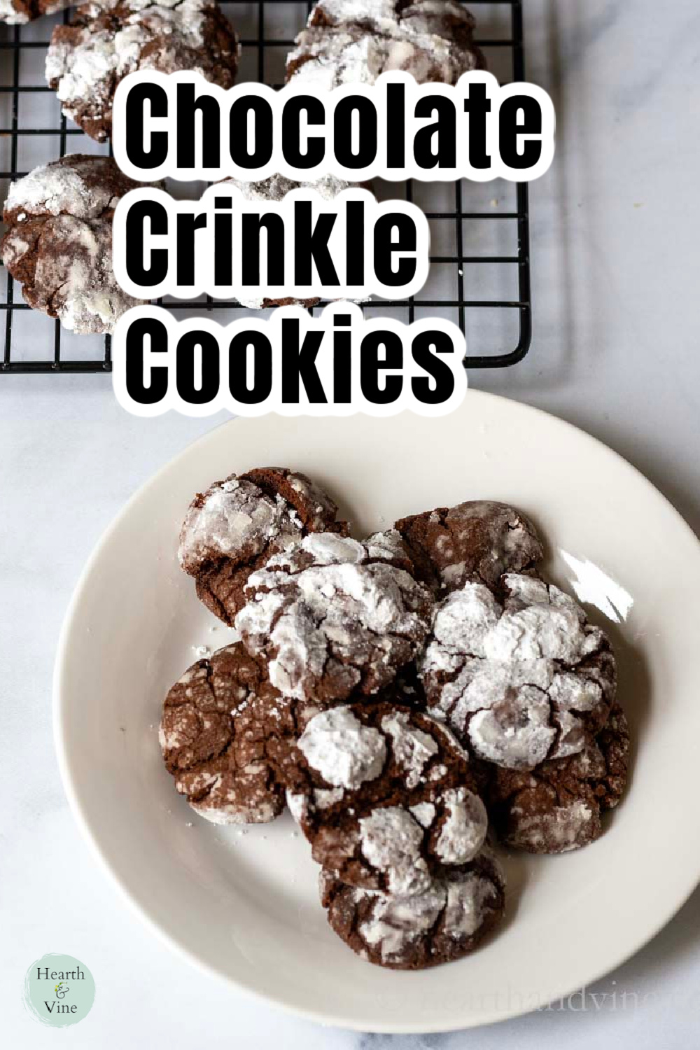 A small white plate of chocolate crinkle cookies below a cooling rack of the same cookies.
