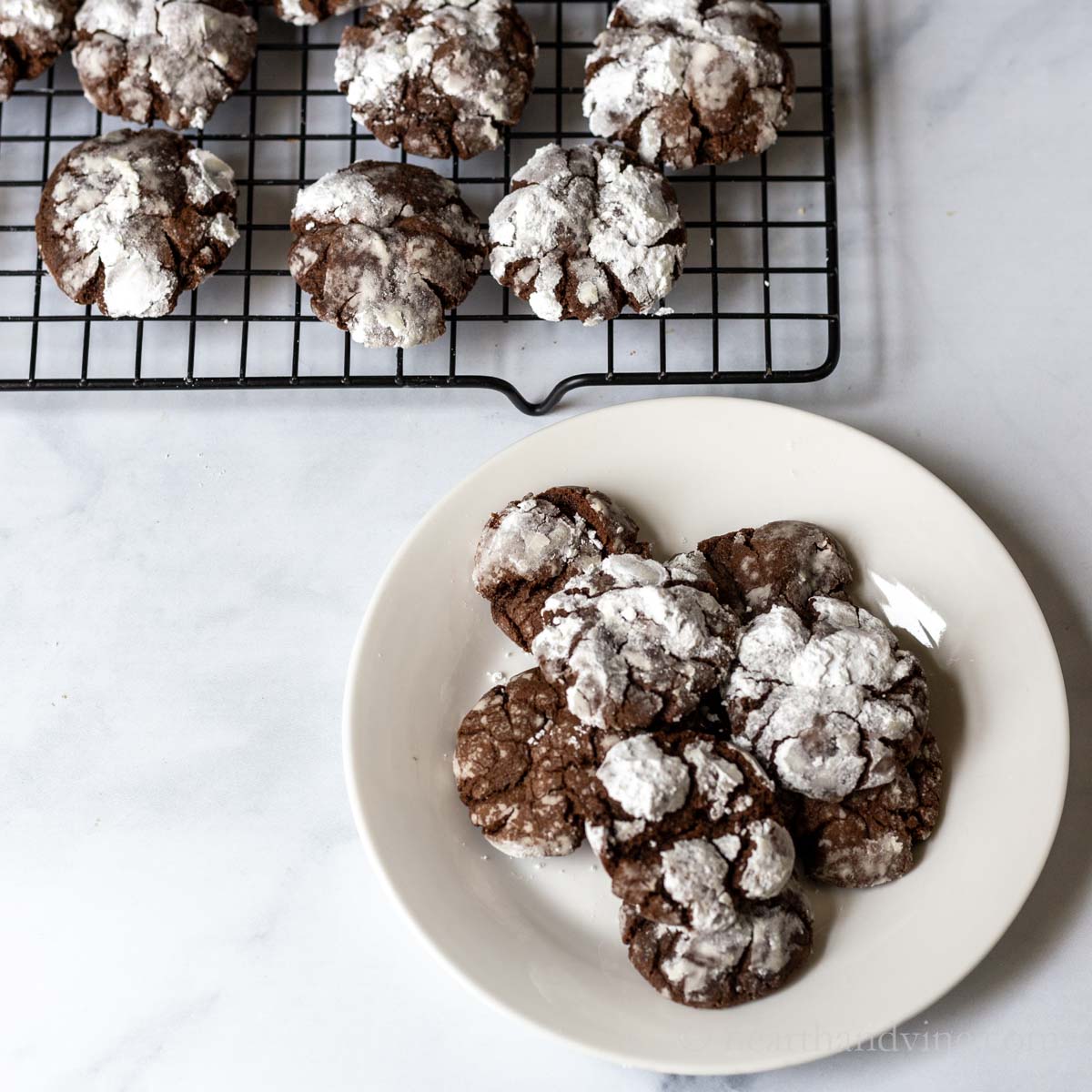 Chocolate crinkle cookies on a white plate with some more of the same on a cooling rack behind.
