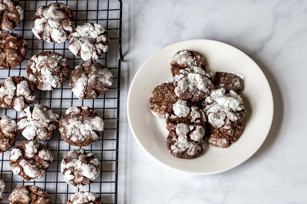 Plate of chocolate crinkle cookies  next to a cooling rack of the same.