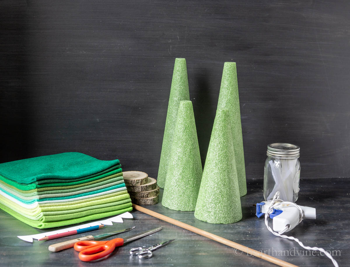 Felt Christmas trees supplies including, a dowel road felt in shades of green, glue sticks a glue gun, floral foam cones, wood slices and scissors.