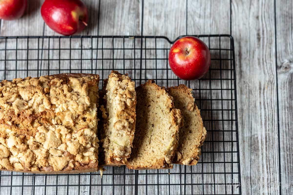 Apple bread partially slice on a wire rack with a few mini red apples on the side.