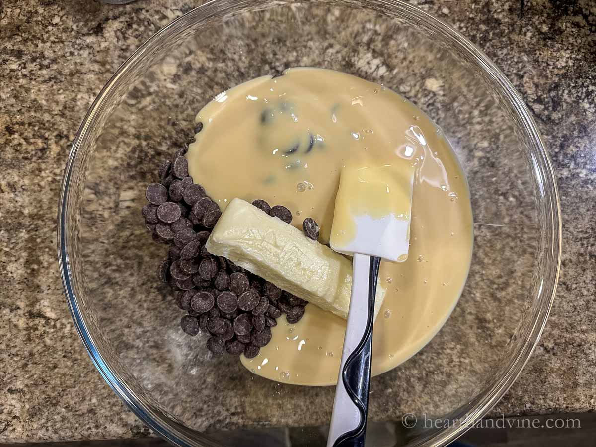 Chocolate chips, a stick of butter, and sweetened condensed milk in a bowl with a rubber spatula.