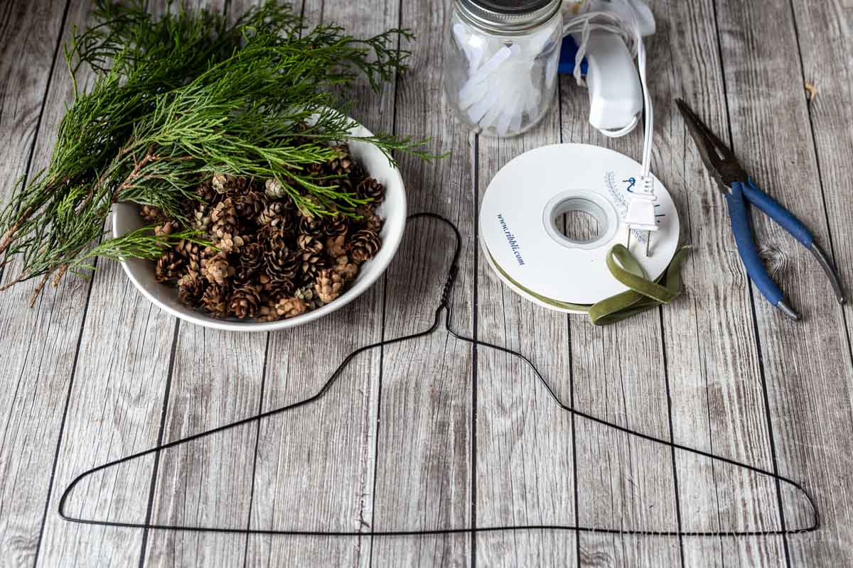 Supplies for mini pinecone wreaths including a wire hanger, pliers, glue gun, glue sticks, mini pinecones, and cedar branches.
