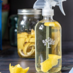 Large spray bottle with lemon vinegar cleaner a few lemon peels scattered and a jar of lemon peels in the background.
