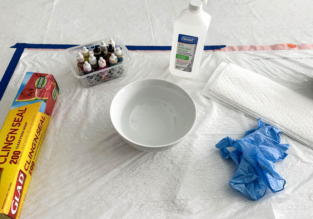 Supplies for alcohol ink with plastic craft including inks, rubbing alcohol, plastic wrap, ceramic bowl and disposable gloves.