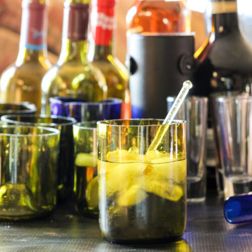A small glass made from a wine bottle with ice and a stirrer.