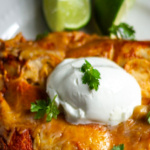 A close up look at two chicken enchiladas on a plate with a dollop of sour cream and some cilantro. A couple of lime wedges on the side.