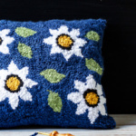 Deep blue punch needle pillow with white daisy flowers and green leaves.