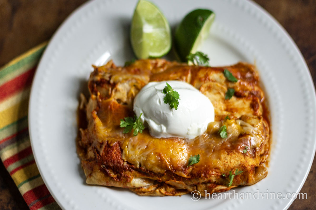 Two chicken enchiladas on a plate with a dollop of sour cream and some cilantro. A couple of lime wedges on the side.