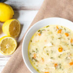 Serving of lemon chicken orzo soup in a white bowl next to a few cut lemons.
